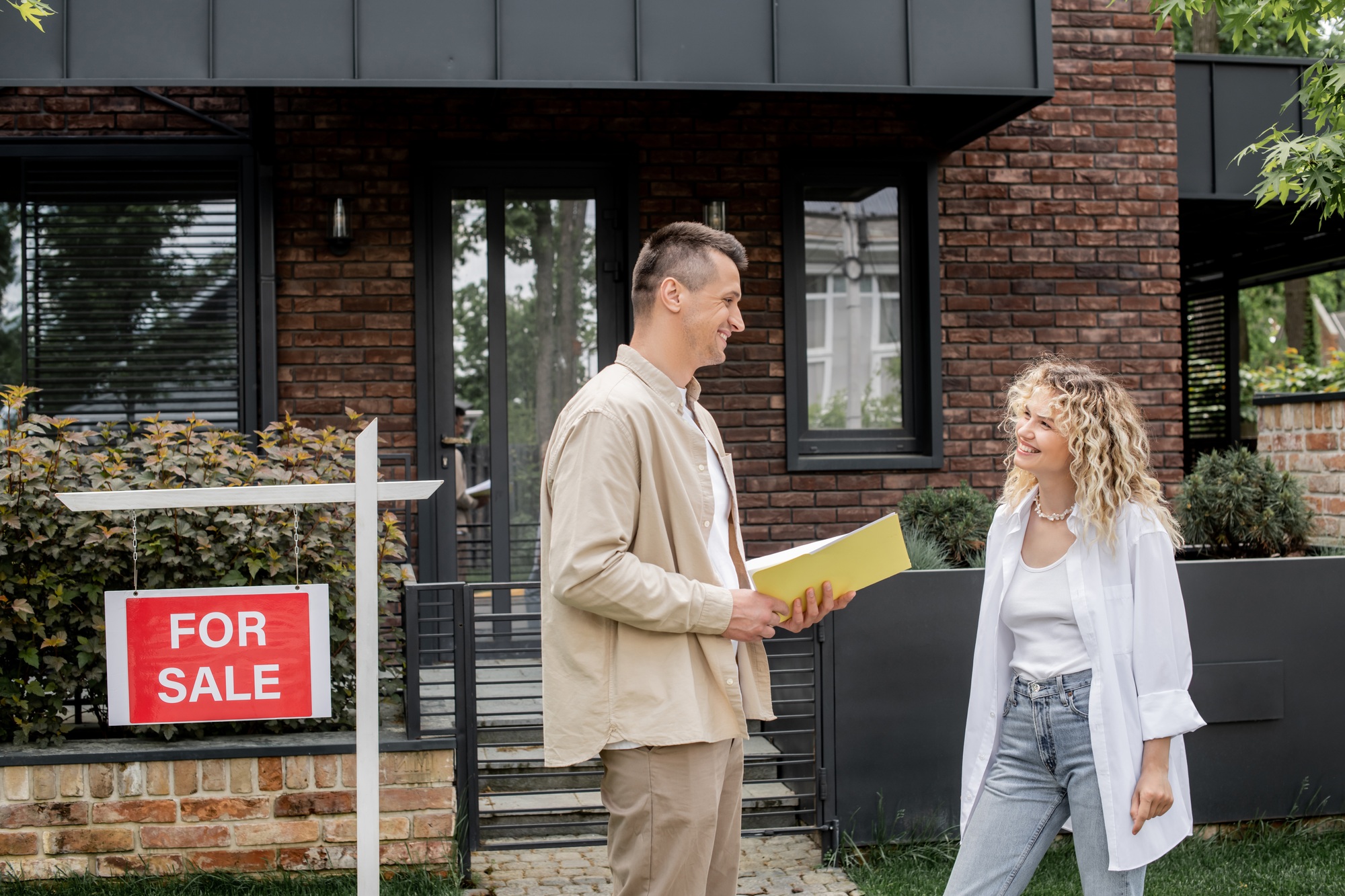 cheerful property realtor with folder talking to woman near modern cottage in city