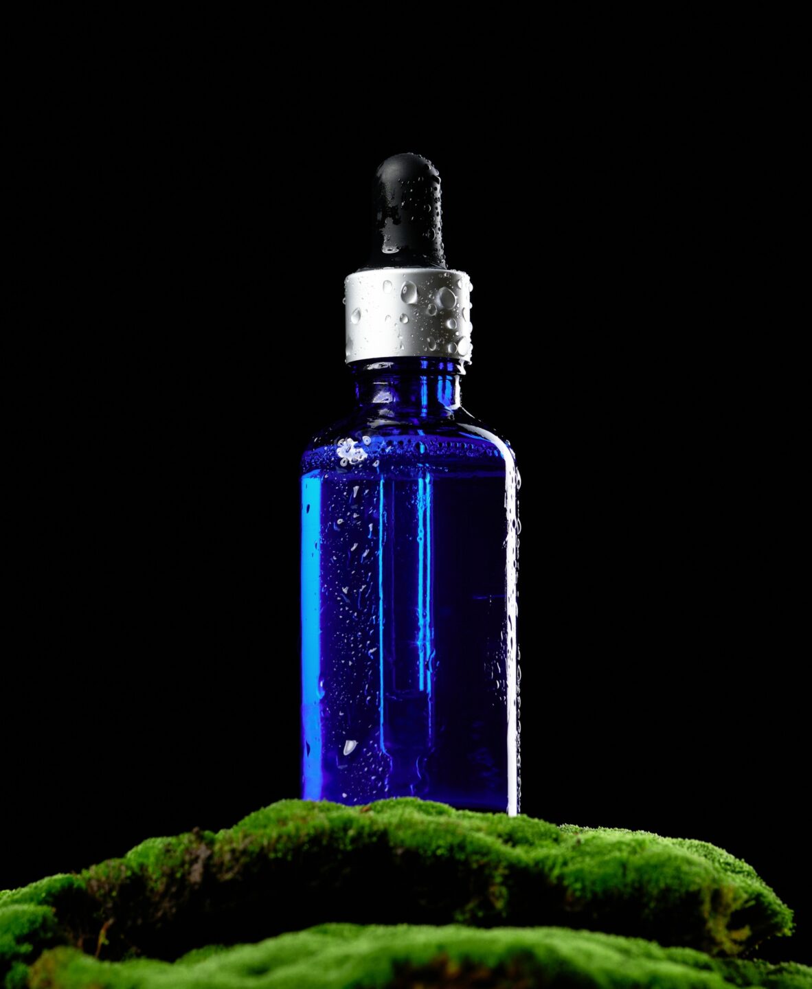 Blue glass bottle with pipette stands on green moss, black background. Cosmetics SPA branding mockup