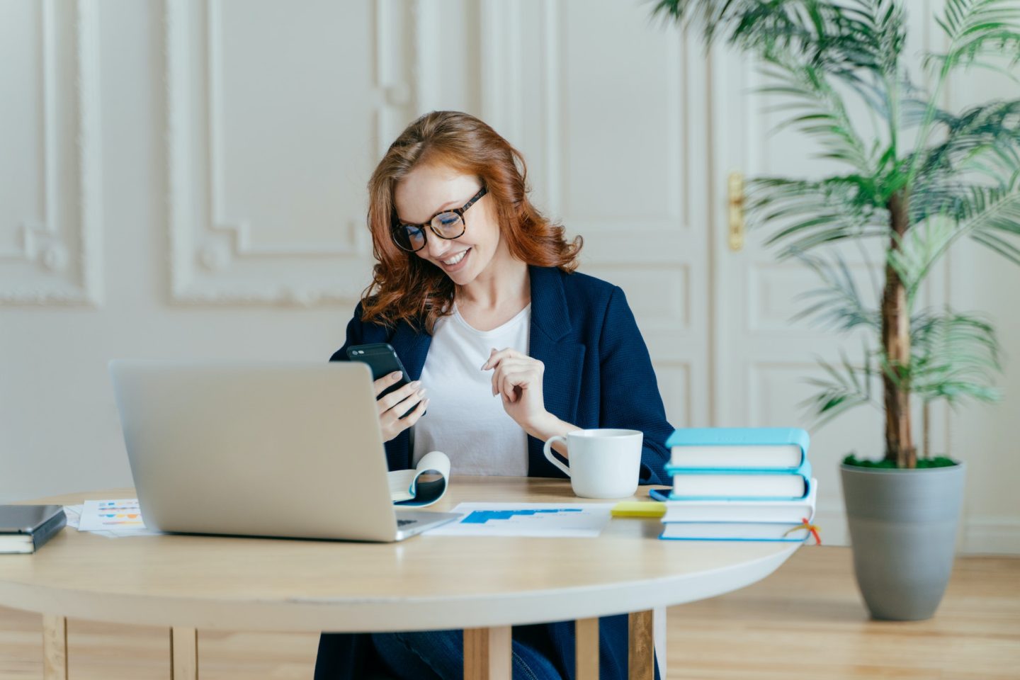 Female focused in modern cellular, happy to receive text message, poses at workplace, drinks coffee.