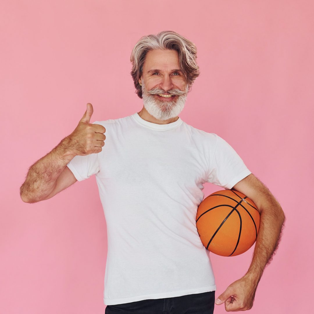 With sport ball in hands. Stylish modern senior man with gray hair and beard is indoors