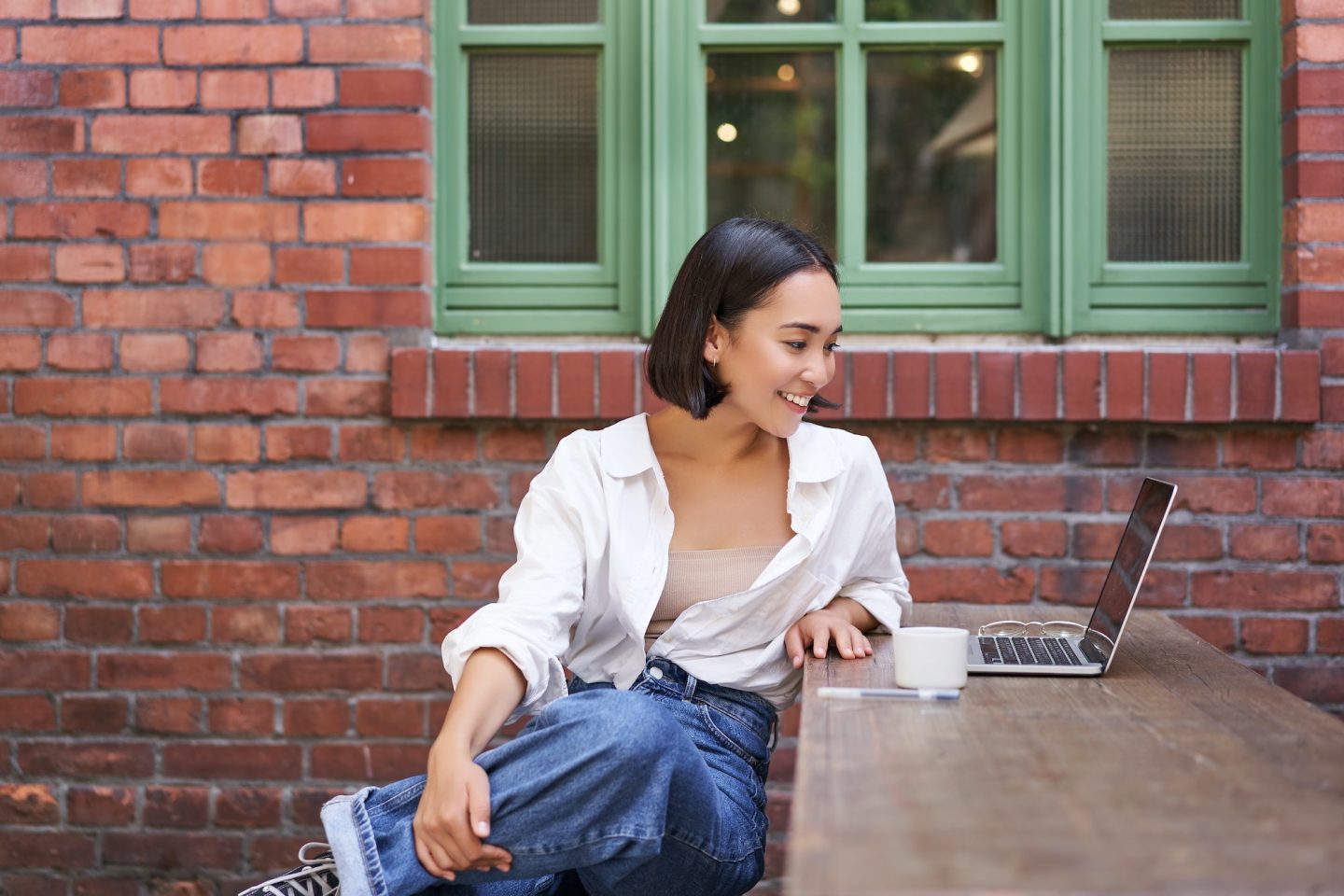 Portrait of young stylish woman, influencer sitting in cafe with cup of coffee and laptop, smiling
