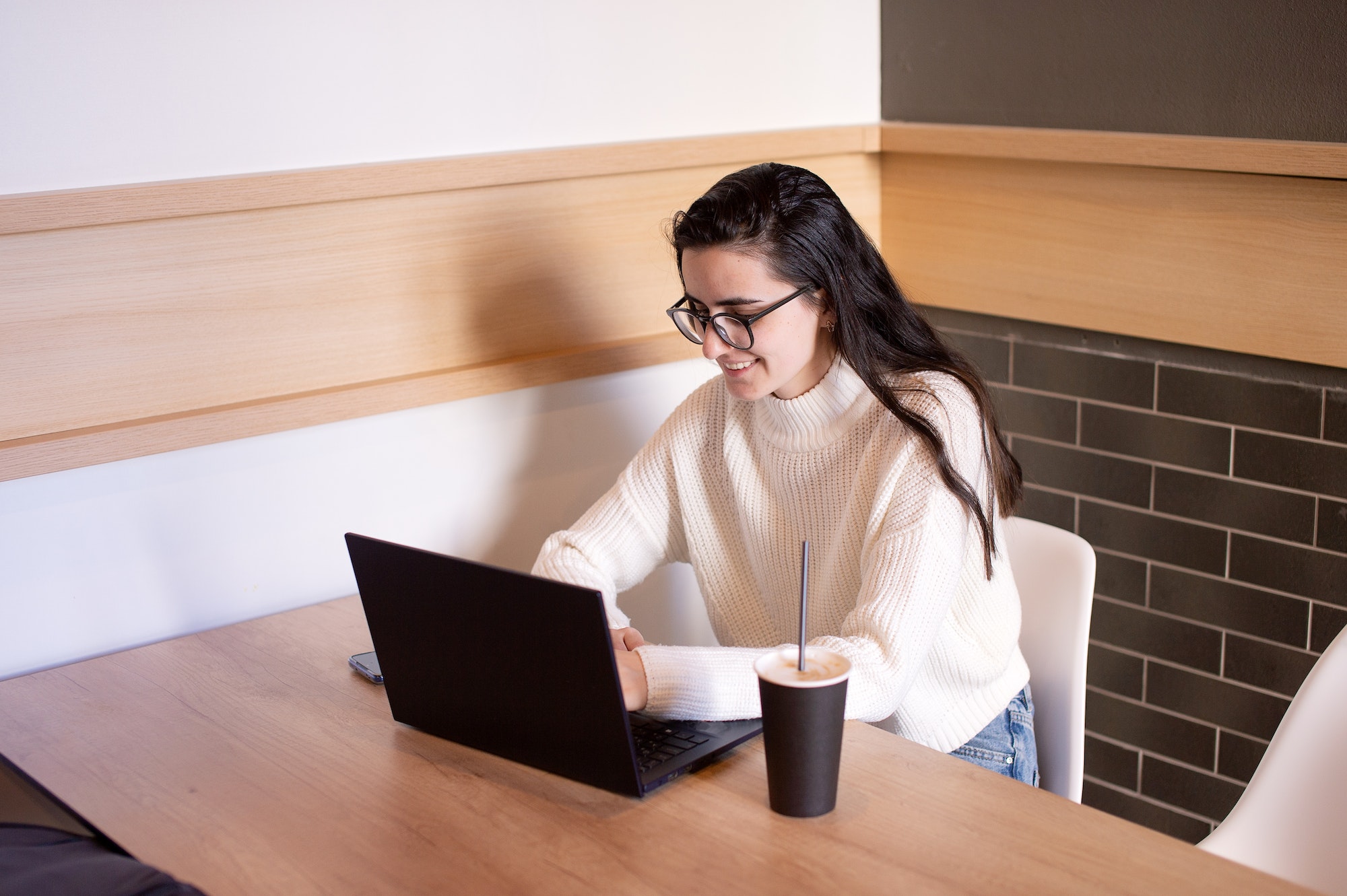 A young girl is sitting in a cafe with a phone and laptop and working